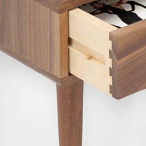 May include: A wooden end table with a drawer that is open, revealing a magazine inside. The table has a walnut finish and four legs.