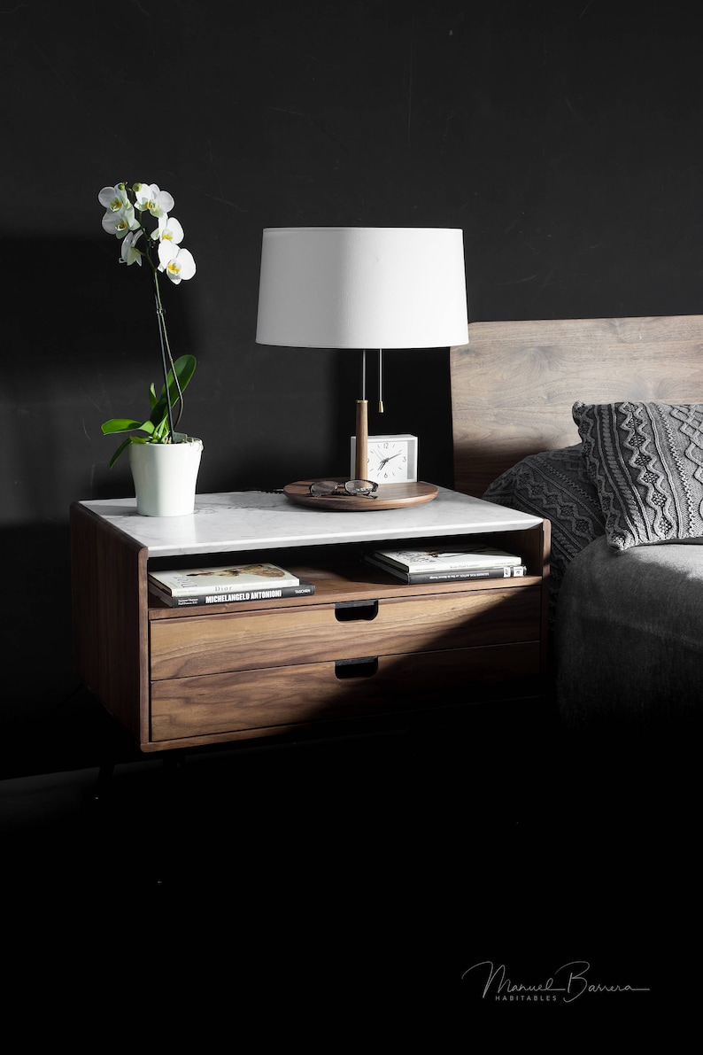 May include: A wooden nightstand with two drawers and a white marble top. A white lamp with a white shade sits on the top of the nightstand, along with a clock and a pair of glasses. A white orchid plant is in a white pot on the nightstand. The nightstand is next to a bed with a gray knitted throw pillow.