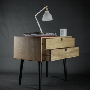 May include: A wooden nightstand with two drawers and black legs. The top drawer is open and a book titled "The Man of Fashion" is on top of the nightstand.