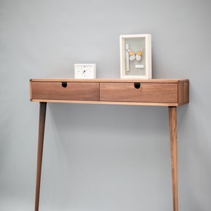 May include: A walnut wood console table with two drawers and tapered legs. The table has a white clock and a framed butterfly specimen on top.