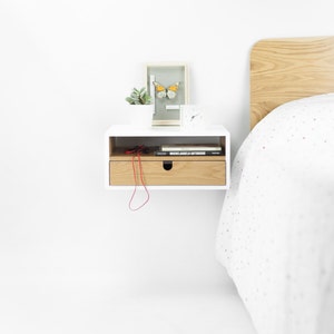 May include: White floating nightstand with a drawer, a small potted plant, a framed butterfly, and a clock on top. The drawer is open and has a red cord and two books inside. The nightstand is mounted on a white wall next to a bed with a white and gray patterned duvet.