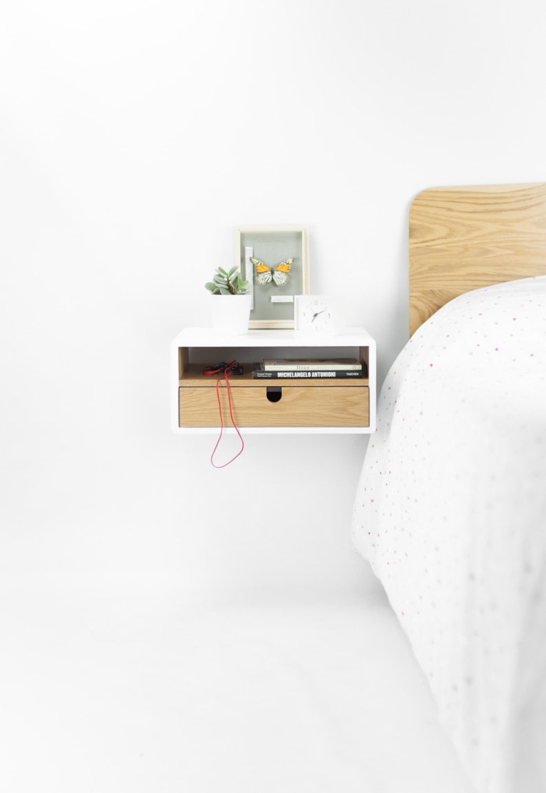 May include: White floating nightstand with a drawer, a small potted plant, a framed butterfly, and a clock on top. The drawer is open and has a red cord and two books inside. The nightstand is mounted on a white wall next to a bed with a white and gray patterned duvet.
