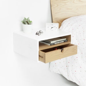 May include: White floating nightstand with a drawer and a shelf. The nightstand is mounted to a white wall and has a small potted plant, a clock, and glasses on top. The drawer is made of light wood and is open.