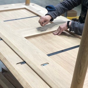 May include: A wooden table top being assembled with black metal brackets and wooden legs. The table top is made of light wood and has a natural finish.