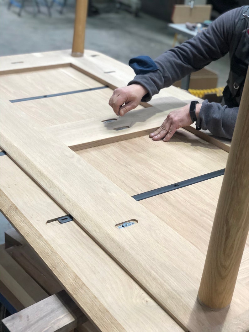 May include: A wooden table top being assembled with black metal brackets and wooden legs. The table top is made of light wood and has a natural finish.