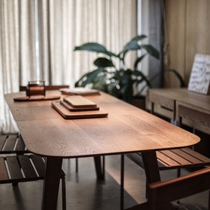 May include: A wooden table with a light wood finish and a rounded edge. The table has four legs and is set with three wooden cutting boards. Two wooden chairs are visible in the foreground.