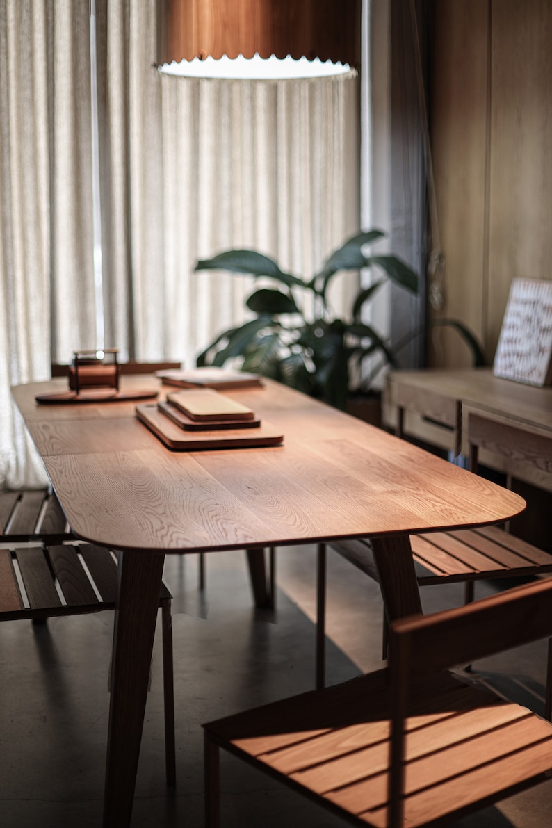 May include: A wooden table with a light wood finish and a rounded edge. The table has four legs and is set with three wooden cutting boards. Two wooden chairs are visible in the foreground.