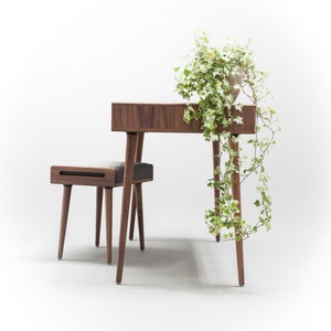 May include: A wooden desk with a light wood finish and a small stool. The desk has a drawer and four legs. The stool has a gray fabric seat and four legs. A potted plant with green leaves hangs over the desk.