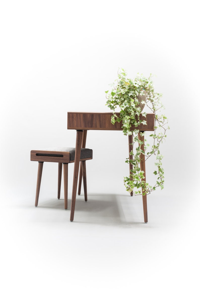 May include: A wooden desk with a light wood finish and a small stool. The desk has a drawer and four legs. The stool has a gray fabric seat and four legs. A potted plant with green leaves hangs over the desk.
