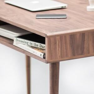 May include: A walnut wood desk with a shelf underneath. The shelf holds a white book with the title "Dressing in the Dark" visible. The desk has a laptop, a phone, and a small plant on top.