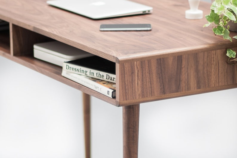 May include: A walnut wood desk with a shelf underneath. The shelf holds a white book with the title "Dressing in the Dark" visible. The desk has a laptop, a phone, and a small plant on top.
