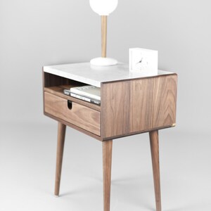 May include: A walnut wood nightstand with a white marble top. The nightstand has a drawer and a shelf. It has four legs and a white lamp and clock on top.