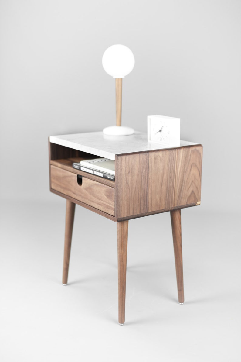 May include: A walnut wood nightstand with a white marble top. The nightstand has a drawer and a shelf. It has four legs and a white lamp and clock on top.