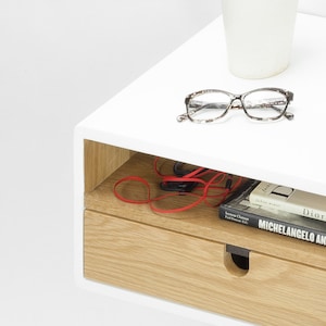May include: White floating shelf with a drawer and a small open shelf. The shelf has a potted succulent, glasses, and headphones on it. The drawer is made of wood and has a cutout handle. The books on the shelf are "Fashion Memoir" by Iris Apfel, "Seymour Chwast" by Paul Duncan, and "Michelangelo and the Sistine Chapel" by Antonio Paolucci.
