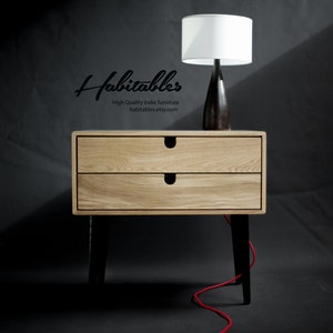 May include: A light wood nightstand with two drawers and black legs. A white lamp with a black base sits on top of the nightstand. A red cord is on the floor.