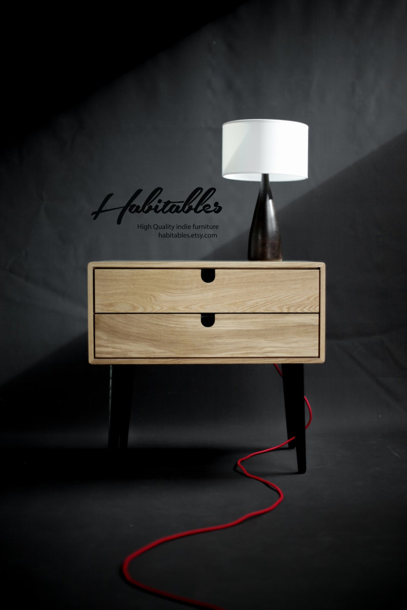 May include: A light wood nightstand with two drawers and black legs. A white lamp with a black base sits on top of the nightstand. A red cord is on the floor.