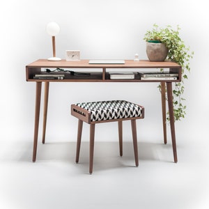 May include: A wooden desk with a shelf and a small stool. The desk has a lamp, a potted plant, and a laptop on it. The stool has a black and white patterned cushion.