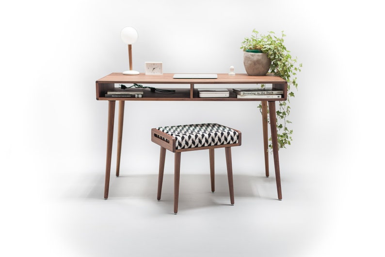 May include: A wooden desk with a shelf and a small stool. The desk has a lamp, a potted plant, and a laptop on it. The stool has a black and white patterned cushion.