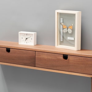 May include: A walnut wood console table with two drawers and black metal drawer pulls. The table has a white clock and a framed butterfly specimen on top.