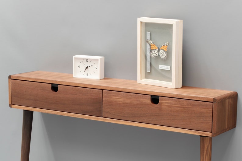 May include: A walnut wood console table with two drawers and black metal drawer pulls. The table has a white clock and a framed butterfly specimen on top.