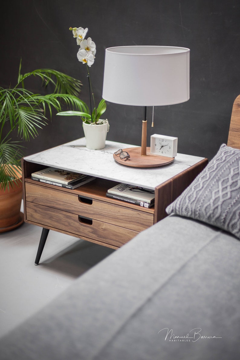 May include: A wooden nightstand with two drawers and a white marble top. A white lamp with a white shade sits on the top of the nightstand, along with a clock and a pair of glasses. A gray throw pillow sits on a bed next to the nightstand.