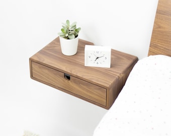 May include: A wooden floating nightstand with a drawer, a small white clock, and a potted succulent plant. The nightstand is mounted to a white wall next to a white bed with a white and pink patterned bedspread.