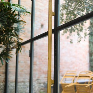 May include: A wooden door handle with a natural finish, mounted on a black metal door frame. The handle is long and slender, with a simple design. The door is made of glass and has a view of a brick wall and some greenery.