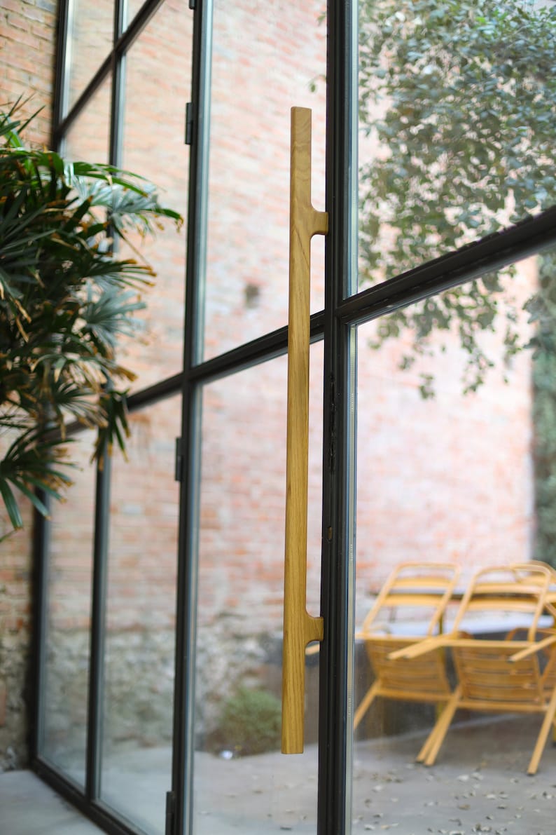May include: A wooden door handle with a natural finish, mounted on a black metal door frame. The handle is long and slender, with a simple design. The door is made of glass and has a view of a brick wall and some greenery.