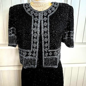 May include: A black cropped jacket featuring intricate silver beadwork in floral and geometric patterns along the front opening, neckline, and sleeve cuffs. The garment is fully covered in small black beads and has short sleeves and a round neckline.
