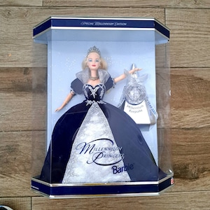 May include: A Barbie doll dressed in a blue and silver gown with a tiara. The doll is in a clear plastic display case with a silver and blue box that says "Millennium Keepsake" and "New Year 2000". The box also says "Special Millennium Edition" and "Millennium Princess Barbie".