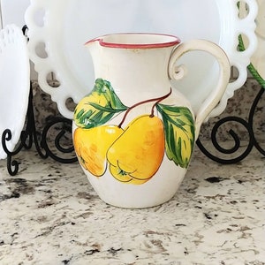 May include: A white ceramic pitcher with a red rim and a handle. The pitcher is decorated with a hand-painted design of two yellow pears with green leaves.