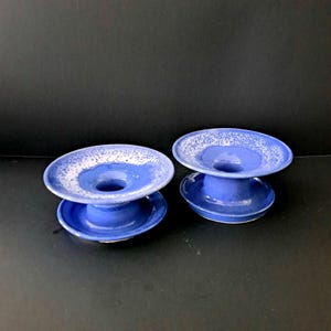 May include: Two blue ceramic candle holders with white speckles. The holders have a wide base and a narrow top with a hole in the center for a candle.
