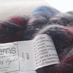 May include: A ball of Artyarns Rhapsody light yarn in shades of gray, burgundy, and blue. The label reads "Artyarns, Elevate your art, Rhapsody light, 85% Silk 15% Kid Mohair, 366m/400yd, Color 907".