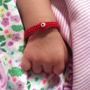 May include: A red string bracelet with a blue evil eye charm on a baby's wrist.