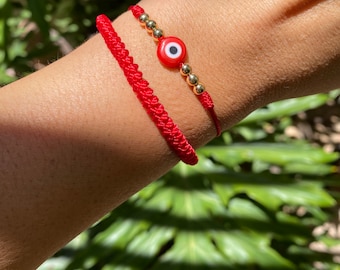 Könnte beinhalten: Zwei rote Armbänder aus Schnur mit goldenen Perlen und einem roten bösen Blick-Anhänger. Die Armbänder werden am Handgelenk getragen.