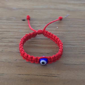 May include: Red string bracelet with a blue evil eye charm. The bracelet is tied with a knot and has a tassel on each end.