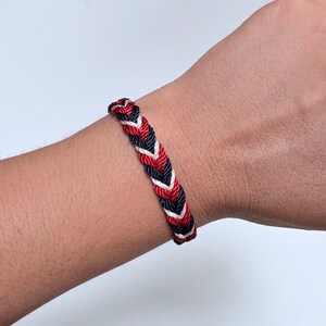 Puede incluir: Una pulsera trenzada que presenta un patrón repetitivo de hilos de cordón retorcidos en azul marino, blanco y rojo. La pulsera está construida con tres colores distintos de cuerda tejidos juntos en un diseño de trenza apretado y uniforme.