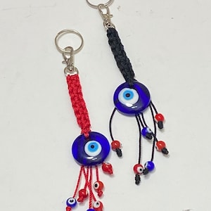 May include: Two blue evil eye keychains with red and black tassels. The keychains are made of braided string and have a silver key ring.