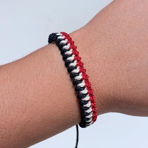 Puede incluir: Una pulsera de cordón trenzado con un patrón repetitivo de hilos negros, blancos y rojos. El diseño consiste en hebras estrechamente anudadas dispuestas en una secuencia diagonal, creando una superficie texturizada. Un cordón negro cuelga.