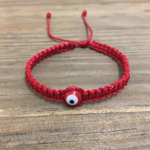 May include: Red string bracelet with a blue evil eye charm. The bracelet is made of braided red string and has a small, round charm with a blue eye in the center.