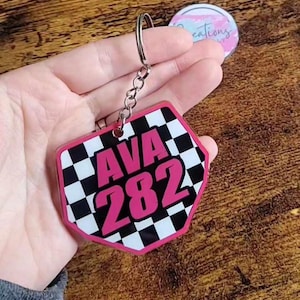May include: A hexagonal keychain featuring a black and white checkered pattern with the name AVA and the number 282 printed in bold pink lettering. The keychain is attached to a silver metal split ring and a short chain link connector.