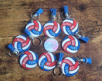 May include: A collection of circular volleyball keychains featuring red, white, and blue panels. Each keychain includes a blue tassel, a metal ring, and custom text including names like Hazel, Maddy, and Payten, jersey numbers, and the acronym EVRC.