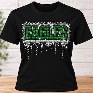 May include: A black short-sleeved t-shirt featuring the word EAGLES in green rhinestones. The text is outlined and accented with white rhinestones that extend downward in a dripping pattern, creating a sparkling effect across the chest area.