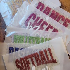 May include: A collection of iron-on rhinestone transfers on clear backing sheets. The designs feature glittery text including "BASKETBALL," "DANCE," "CHEER," "VOLLEYBALL," and "SOFTBALL MOM" in various colours like gold, pink, green, blue, and red.