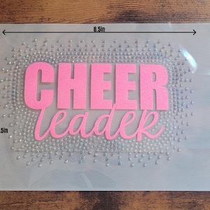 May include: A rectangular iron-on transfer sheet measuring 21.59 cm by 13.97 cm. It features the pink text CHEER leader surrounded by a border of clear, reflective rhinestones on a transparent adhesive backing for fabric application.