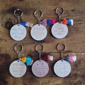 May include: Six wooden keychains with inspirational quotes and colorful tassels. The quotes are: "easy things aren't worth doing", "STOP dreaming START doing", "life is TOUGH but so are YOU", "DREAMS don't work UNLESS you do", "I trust the NEXT CHAPTER because I KNOW THE author", and "do SMALL THINGS with GREAT LOVE".
