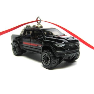 May include: Black toy truck with red stripe detail and large off-road tires. The truck has the word "RAM" on the front and is attached to a red ribbon, suggesting it is an ornament.