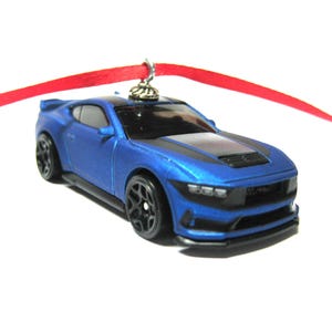 May include: A blue toy car ornament with black accents and black wheels. The car is suspended by a red ribbon, perfect for holiday decorating. The car has a black hood and a detailed front grill.