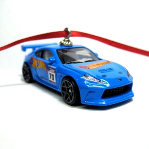 May include: A blue toy car with black wheels and a red ribbon for hanging. The car has the Hot Wheels logo and the number 70 on the side. The car is a miniature replica of a sports car.