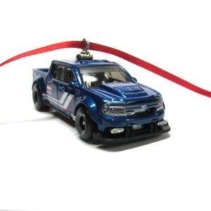 May include: A die-cast blue pickup truck ornament with black wheels and a red ribbon for hanging. The truck has white and blue stripes on the side and a detailed front grill.